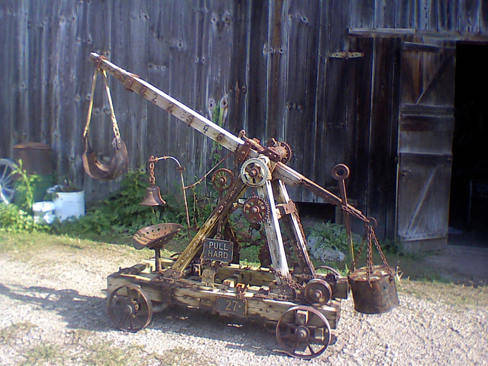 The trebuchet: a throwing machine rebuilt from a burned farm thresher, standing in gravel before a weathered gray barn.